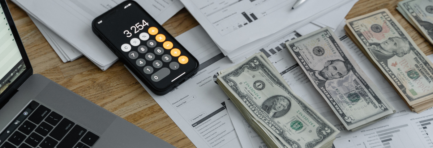 A desk with a laptop, calculator, printed business documents, and organized piles of cash.