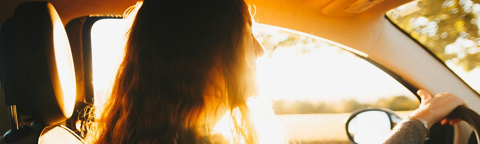 A view from the passenger seat of a young woman driving a car at sunset, lost in thought