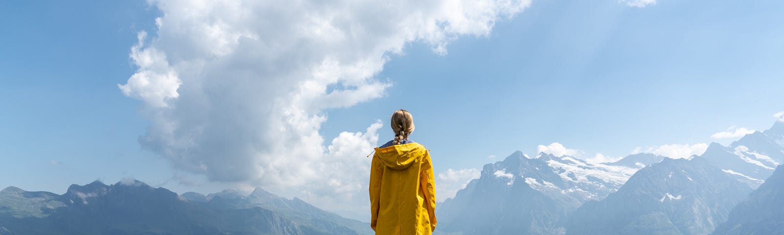 Woman in a yellow coat standing on a hill overlooking mountains.