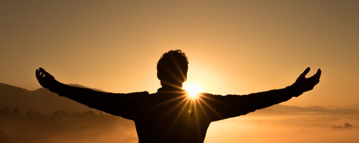 Silhouette of a man standing with outstretched hands and the sun in the background peering over his right shoulder