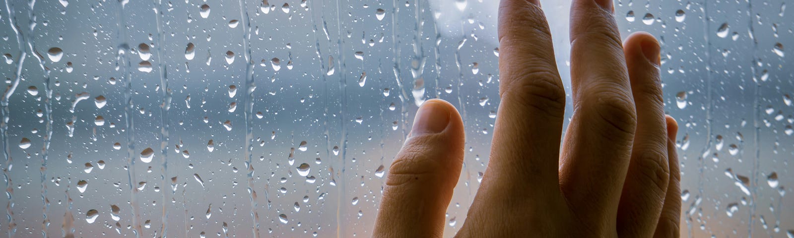 A person’s hand rests against the inside of a window that’s fogged and splattered with rain drops with a view upon an ambiguous landscape of diffused golden colors near the bottom and blue-grey colors near the top, casting an impression of a damp, cloudy morning.