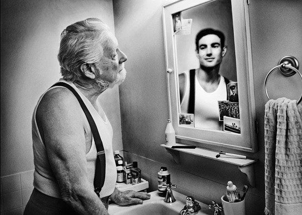 a grey-bearded man in suspenders and an undershirt gazes at a younger version of himself in a mirror