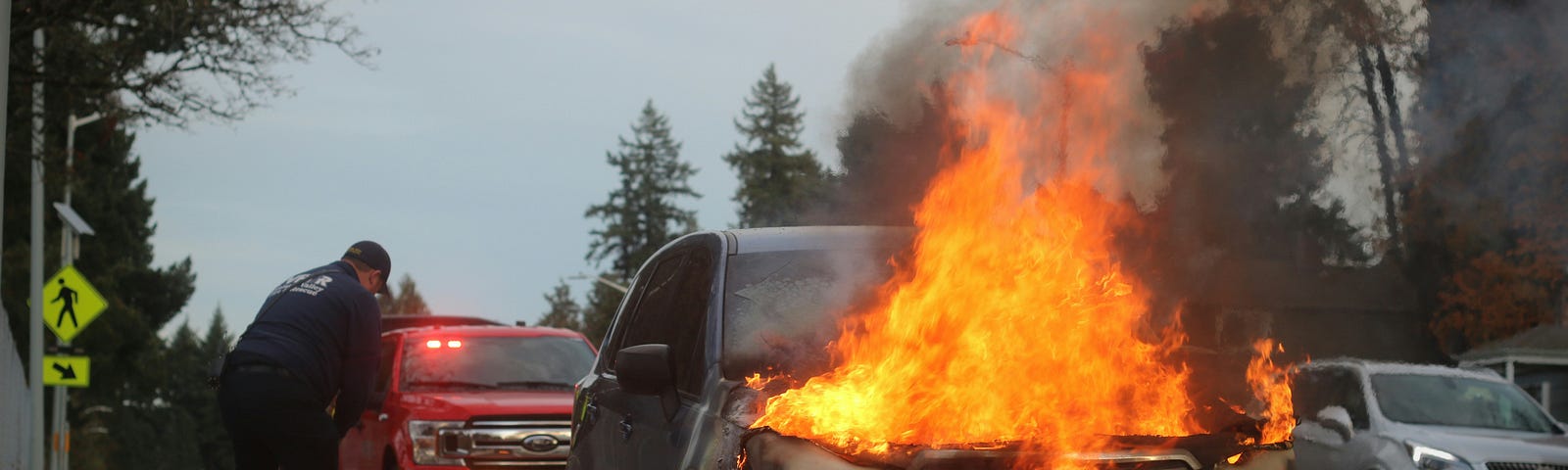 An SUV sits at the side of the road with flames coming out and under the hood. It ain’t great.