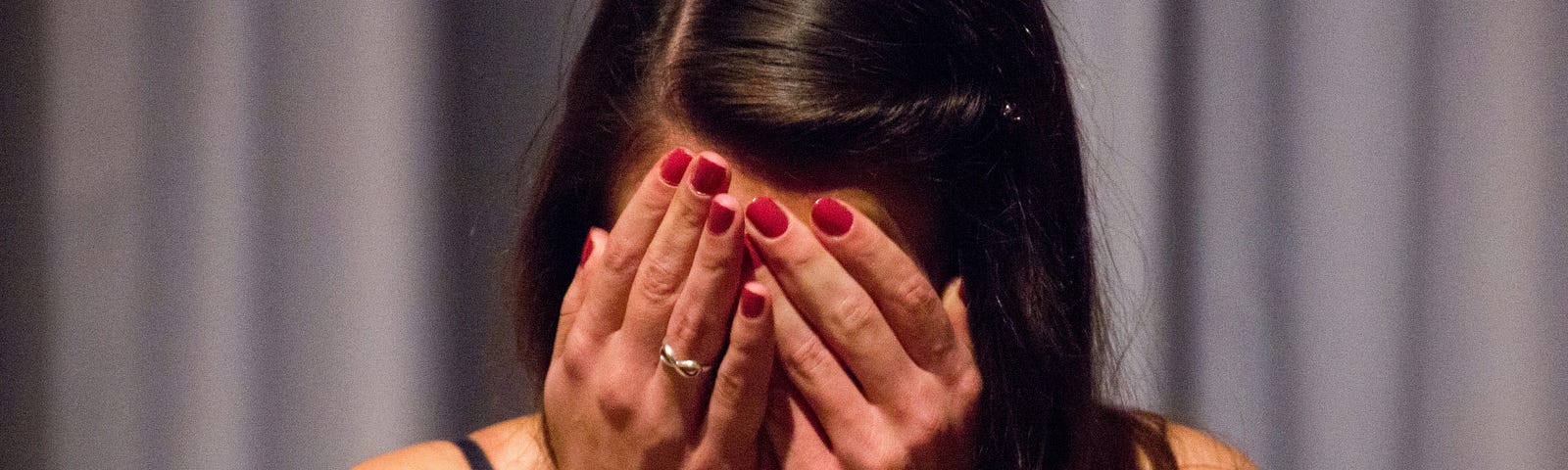 Woman with her head slightly down covering her face with both hands. She has a wedding ring on and red fingernail polish. Her hair is long and dark brown, with a part on the right side.