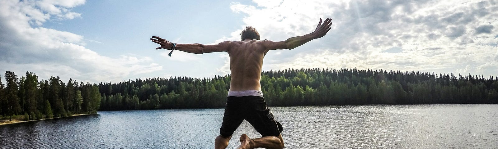 Man with outstretched arms is in mid-jump above the wide expanse of a lake. Blue skies above. The edge of a forest in the distance.