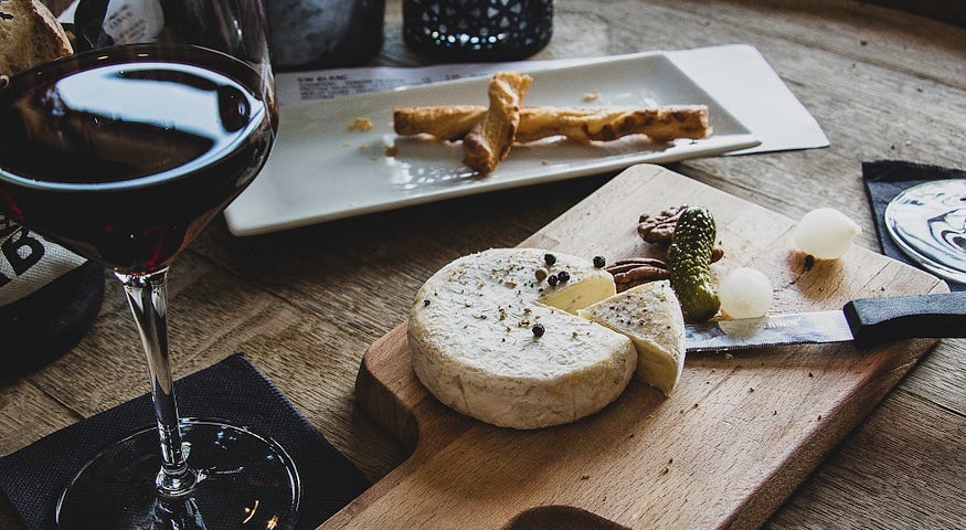 Dark red wine in a tall wine flute on a black napkin next to a wooden cutting board with a cheese wheel, a pie slice cut out, adjacent to a knive and bits of herb and cheese. Behind the cutting board is a ceramic tray with a twisted bread stick on it.