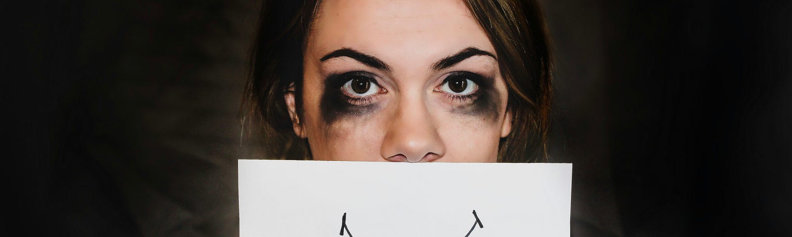 A woman who was crying, with mascara running under her eyes, holding a piece of paper over her face with a smile drawn on it. She has brown hair, a gray-blue shirt, and painted black fingernails