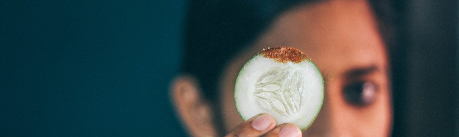 A content-looking woman with brown skin and black hair holds up a teeny slice of cucumber with dip on it.