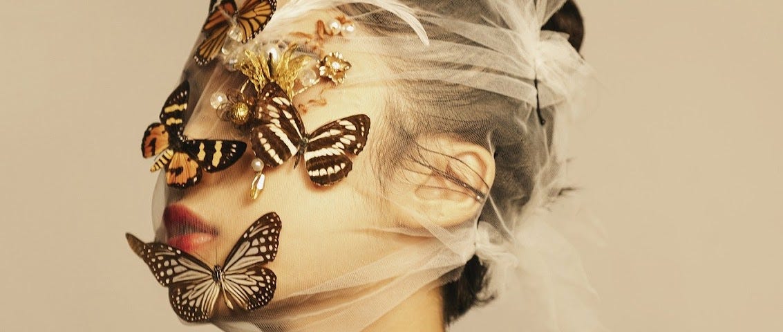 A portrait photo of a light-skinned woman with white gauzy fabric covering her face and five butterflies perched on top of the fabric, obscuring her eyes and nose.