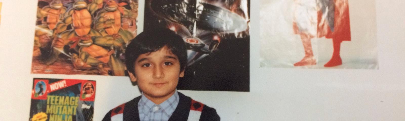 Brown boy around 13 in sweater vest and shirt with Star Trek, Superman Teenage Mutant Ninja Turtles posters behind on wall. Small television on right.