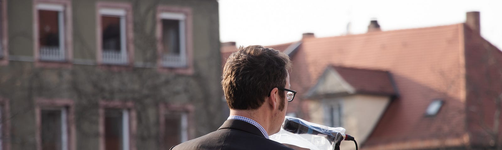 the back of a man in a black suit in the foreground, standing behind a mic, giving a speech, with buildings in the background