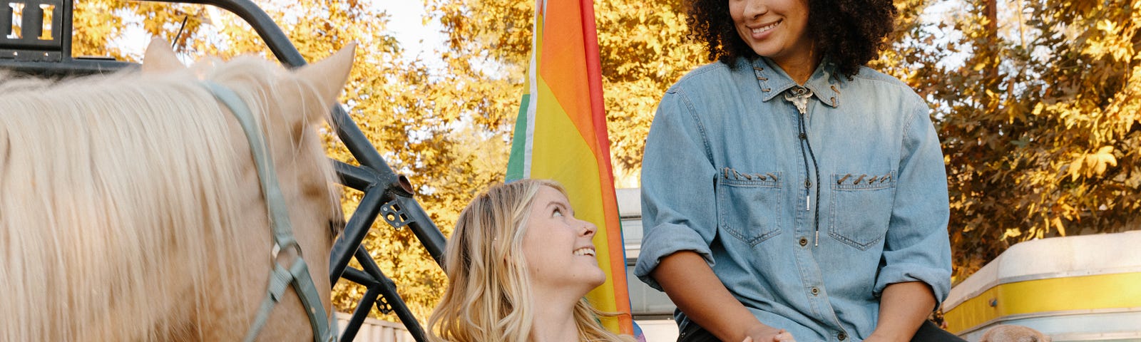 Two women—one Black, one white—holding hands in front of a Pride flag. A horse in a halter is here too.