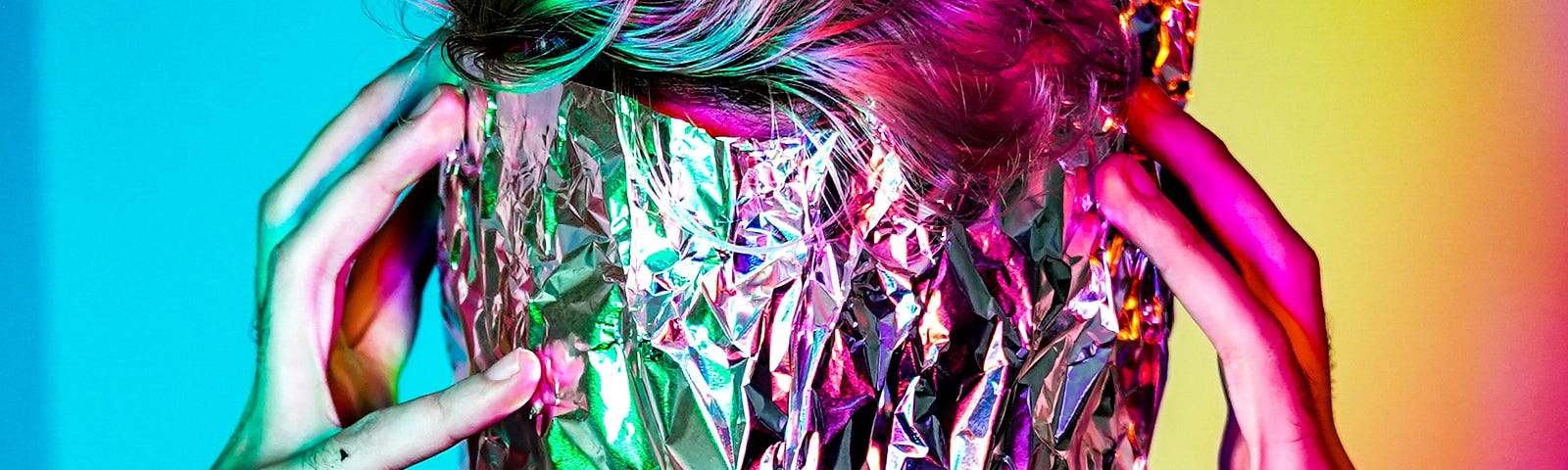 Neon background split evenly into two colors of blue and yellow, with a person squeezing their head, covered in aluminum foil with their finger tips.