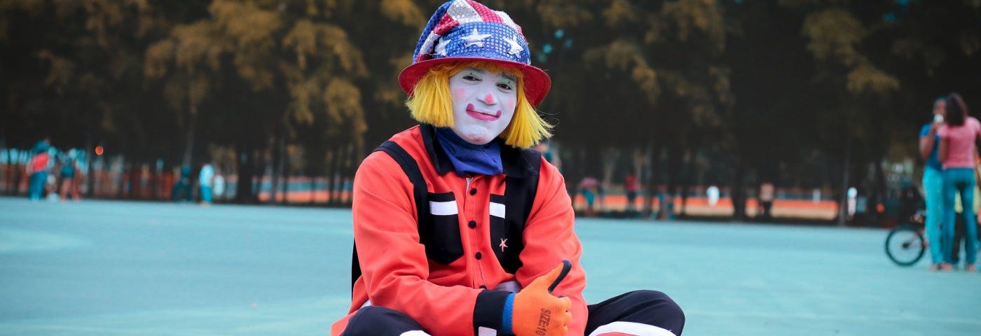 A clown in a stars and stripes hat sits cross legged on the ground.