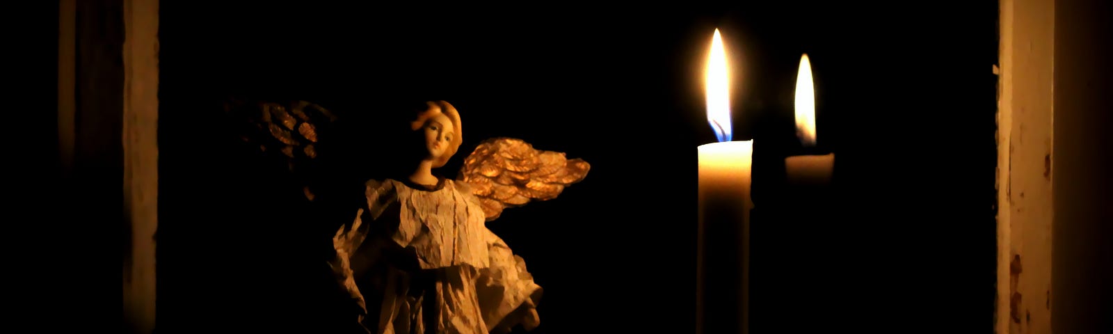 An angel figurine against darkness with two candles casting just enough light to see.