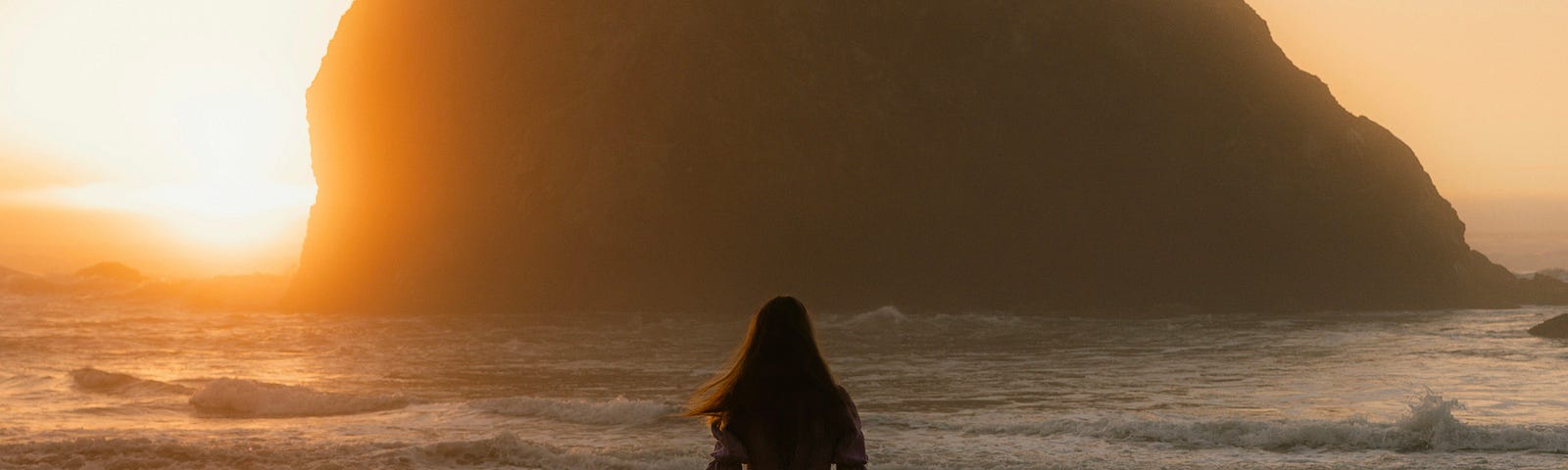 A young woman with long hair, in a dress, sword in hand, looks out on a sunset over the ocean, her back to the viewer.