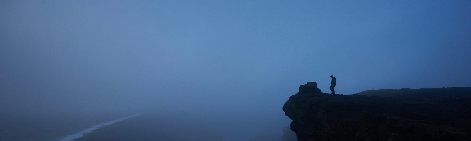 A lone wanderer looks out over a foggy canyon, from a high cliff.