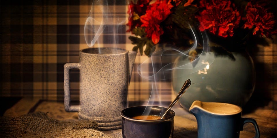 Steaming cup of coffee on a table in a dim-lit image, surrounded by jugs and a vase of red flowers.