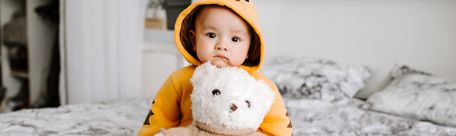 A cute toddler boy sitting on a bed with a white teddy bear in front of him. They boy has a sweet, inquisitive expression.