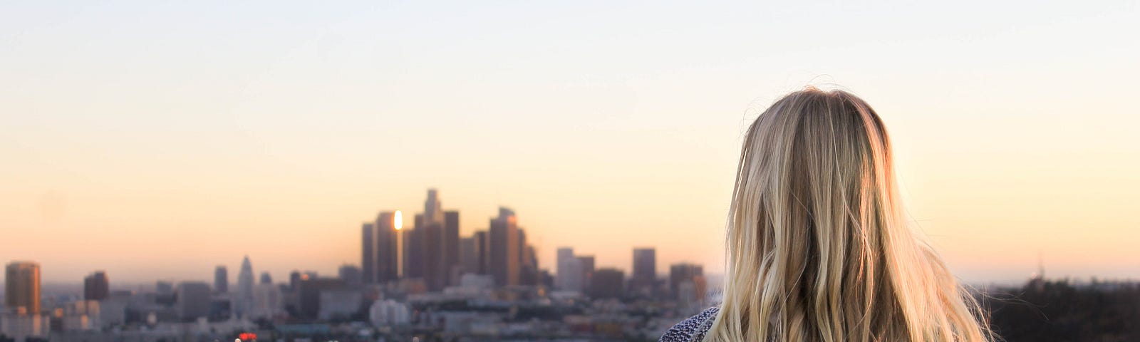 A blonde haired woman, wearing a jumper, stands looking over a cityscape.