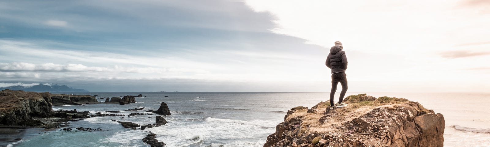 A man standing on the edge of a rock, with a beach, horizon and sky.