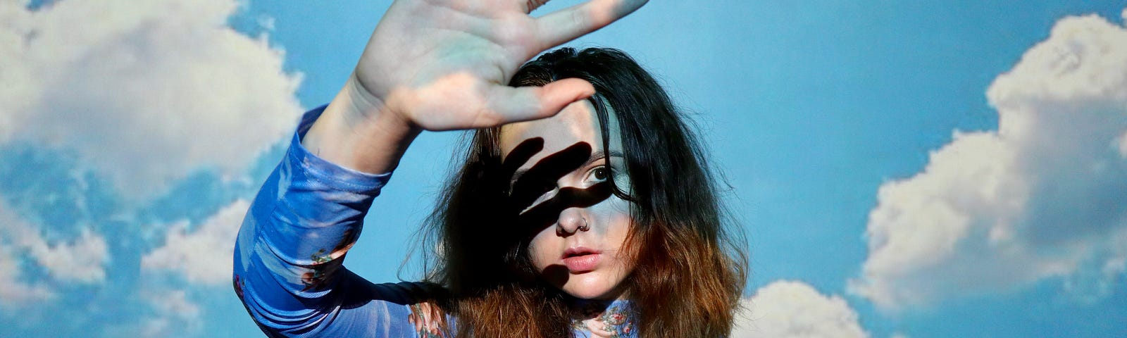 Lady with shoulder-length brown hair, dressed in a print top with long sleeves, holds up her hand, the shadow falling across her face. The background is a blue sky with white clouds.