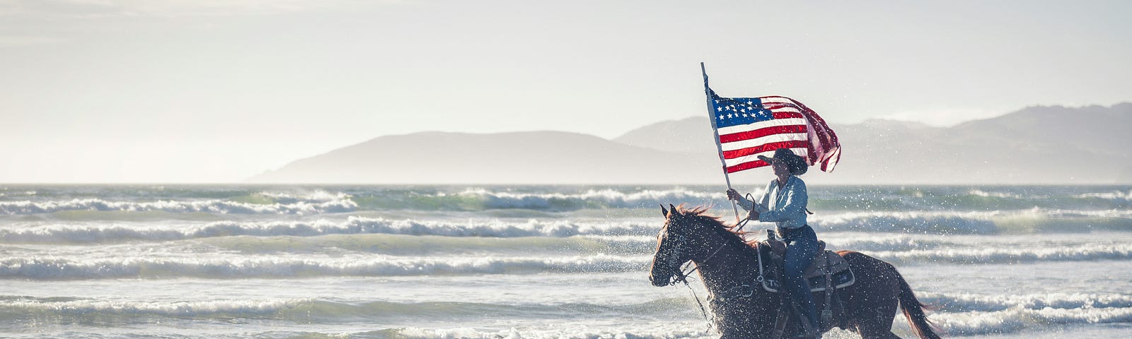 Man on horse galloping through waves while holding an American flag.