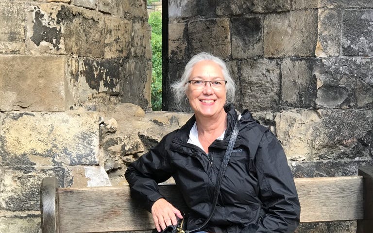 Photo of a gray-haired woman sitting on a wooden bench on front of an old stone building. She is smiling and wearing glasses. She is wearing blue jeans, a black jacket, and black shoes. Her black purse is next to her on the bench.