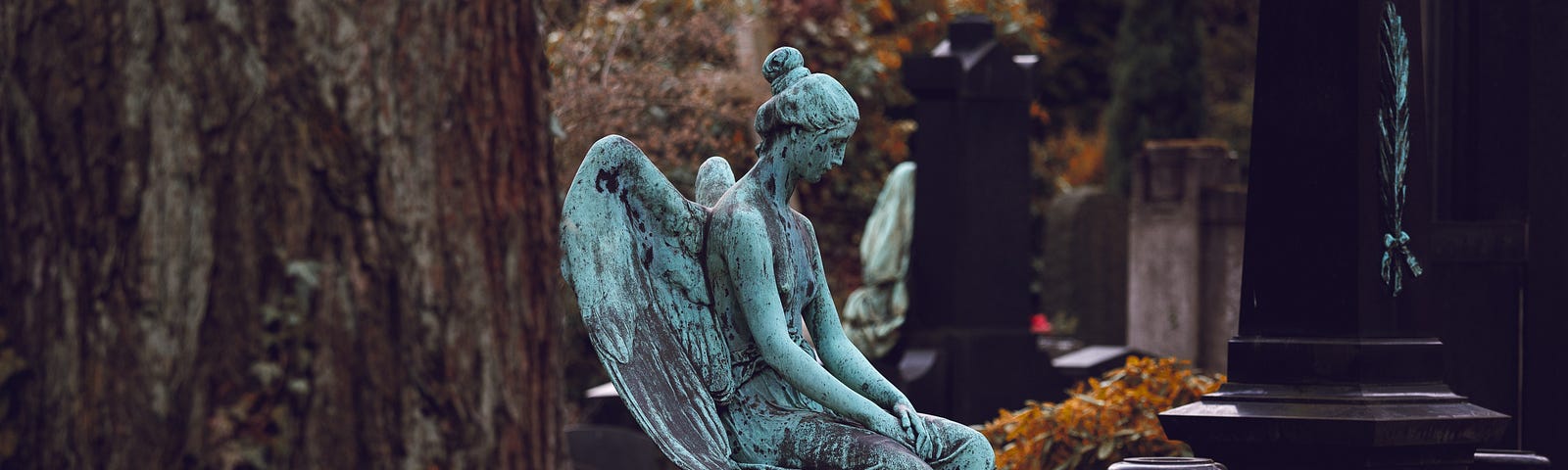 Beyond a tree, a stone angel sits atop a grave looking down at the grave she watches over in a graveyard of black and gray obelisk gravestone.