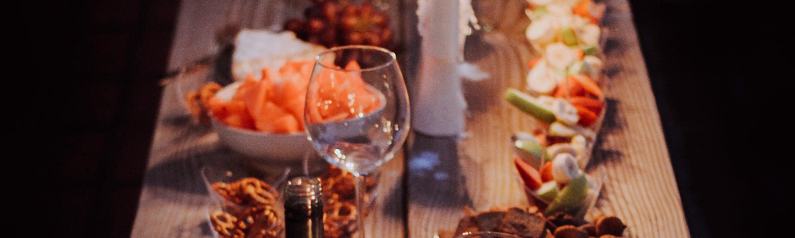 A long wooden board covered in food. A bottle of wine, full glasses, bowls of finger food. The lighting is dim, soft candles against a black background.