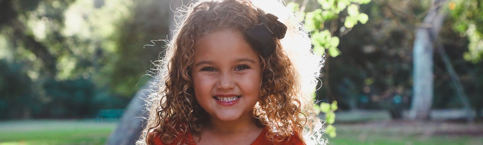 Little girl with long curly hair, wearing a red top, with a big smile.