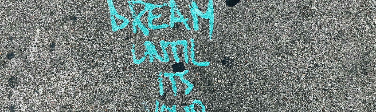 The words, “dream until it’s your reality” printed in turquoise on cement, tips of shoes underneath