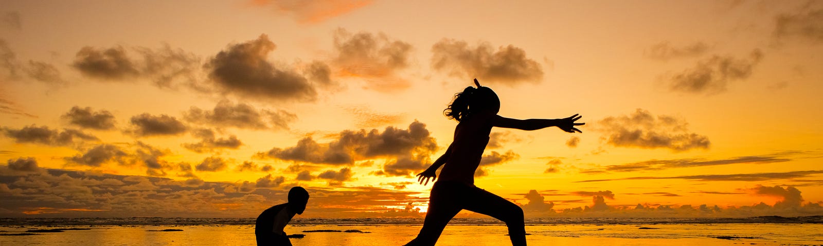 photo of two children playing on the shore, during sunset. The silhouettes of the children show one jumping to the water, and the other standing in the shallow water, with small mounds peaking from the water’s surface, maybe grass, rock, or sand. The sunset behind them is shades of golden yellow, orange and pink, dotted with fluffy clouds.