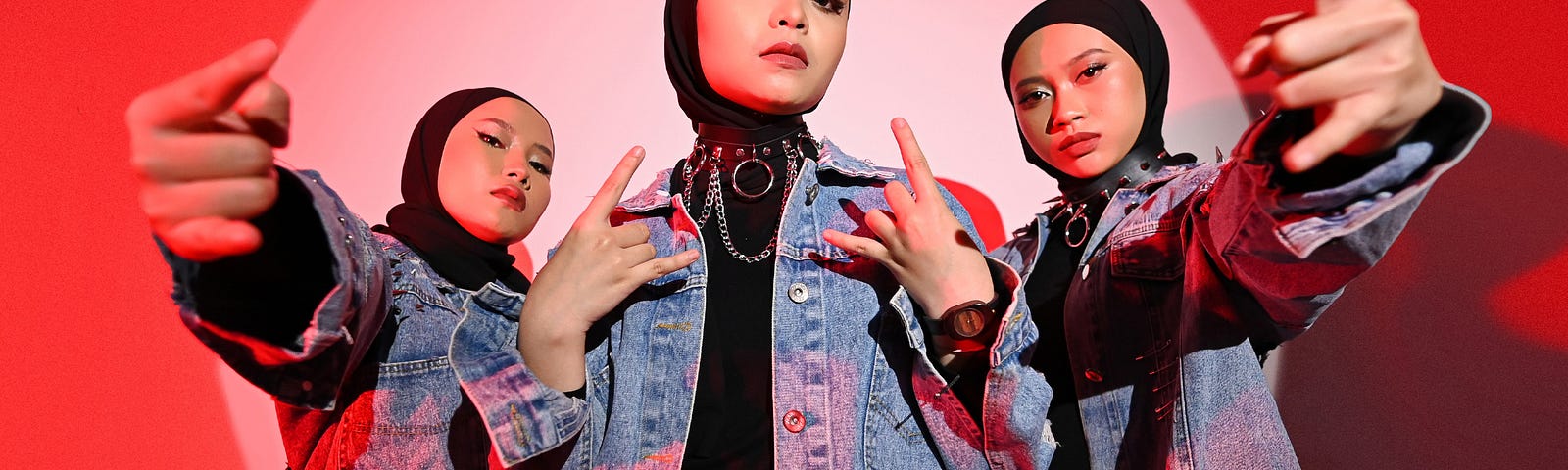 A picture of the three members of Indonesian metal band Voice of Baceprot. The three members are standing in a spotlight against a white backdrop with reddish lighting. All three members are wearing blue denim jackets and pants and black hijabs. All three members have their hands raised in the classic ‘metal’ gesture (a fist, with pinky and pointer raised like a pair of horns).