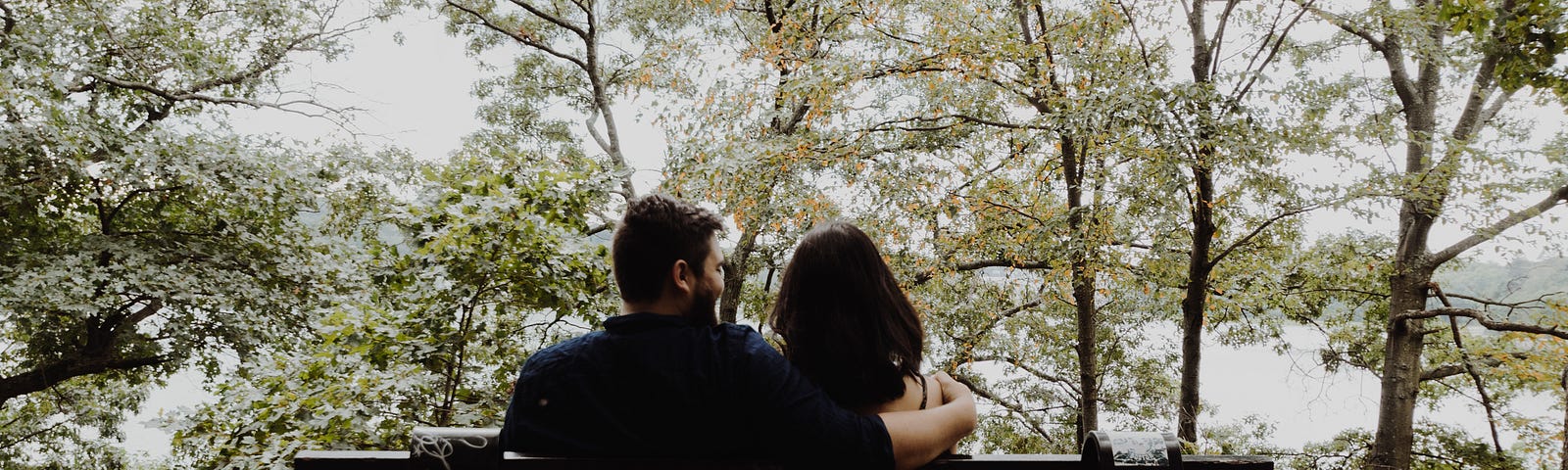 I couple of people sitting on a park bench facing some trees and a lake. One partner has their arm around the other’s shoulders.