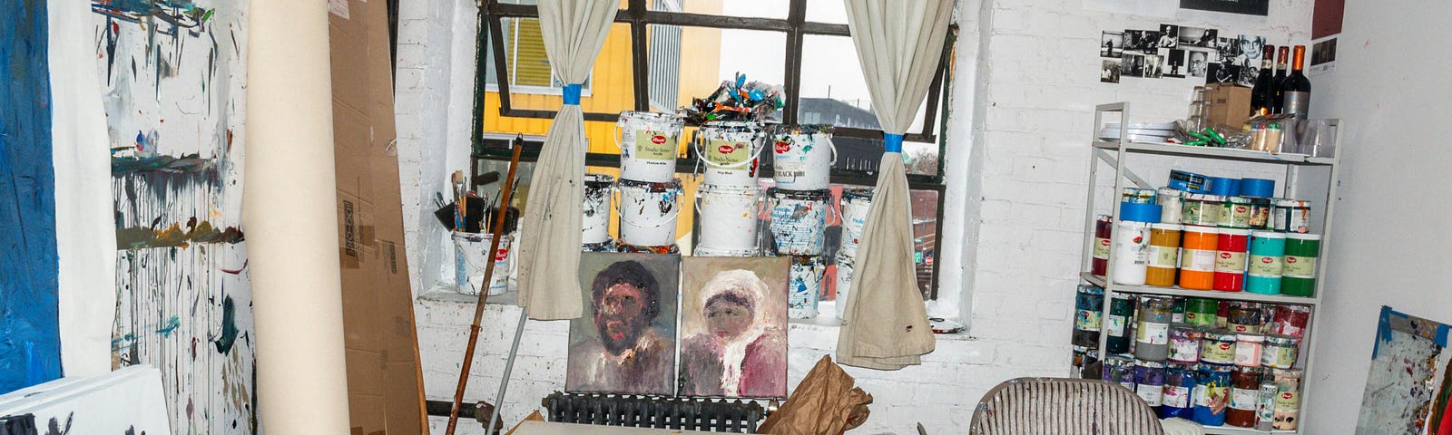 A camera shot of a room with painted canvases leaning on the walls on either side, paint spattered on the floor, cans of paint stacked on the windowsill straight ahead, a shelving unit of paint bottles on the right, and in front of the window, a short table with two portrait paintings, one of a man, and one of a woman.