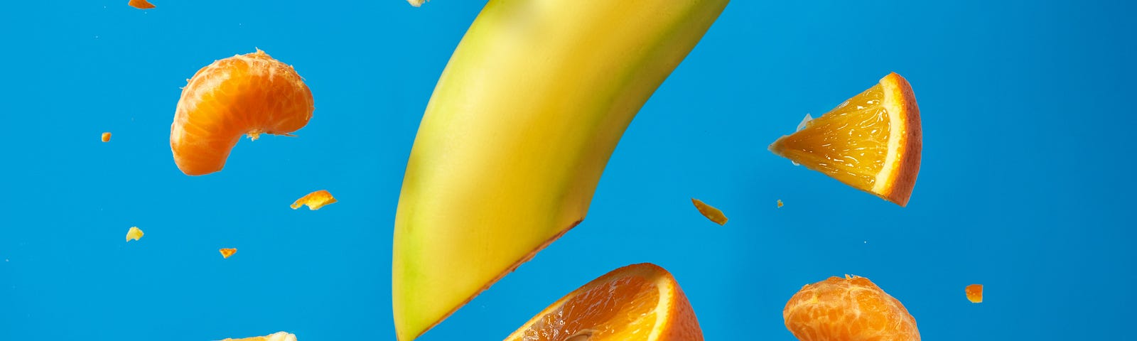 A banana, its end shaped like a dolphin grasping a grape, is surrounded by floating pieces of an orange. Blue background. Cancer remains complex and enigmatic. So many of us have worked for decades to discover effective strategies for cancer prevention.