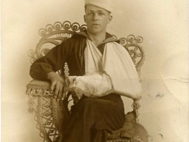 William J. Hughes from Elizabeth City, N.C., sitting on a wicker chair in his U.S. Navy uniform with his arm in a sling.