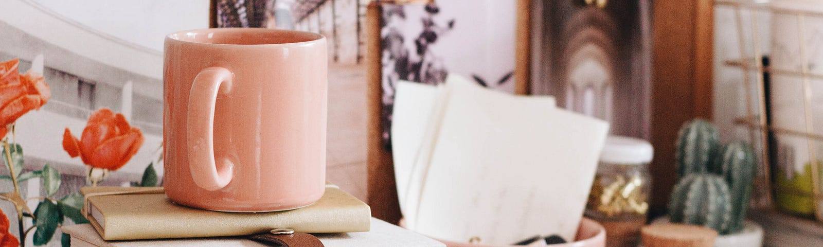 A cluttered, cosy desk space with a stack of novels and notebooks, and a pale pink mug on top.