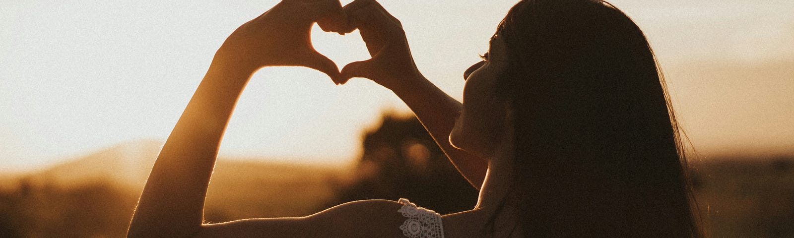 A woman holding her hands in the shape of a heart raised to the sunset.