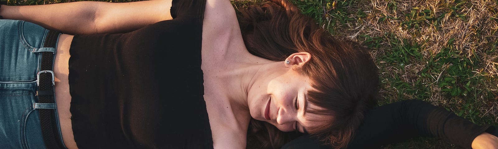 Image of two women who lie on their backs on the grass, their bodies to opposite sides, their heads at each others shoulders and they are looking at each other, smiling.