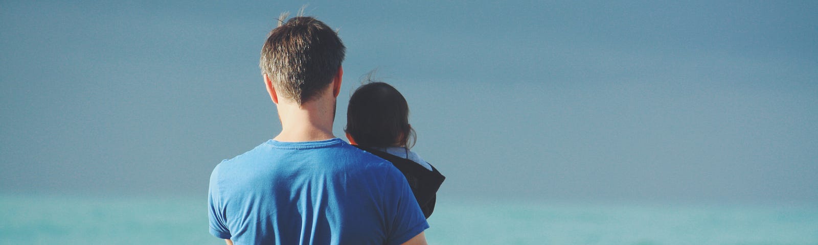 A man holds his small child while facing the view of the ocean waves.