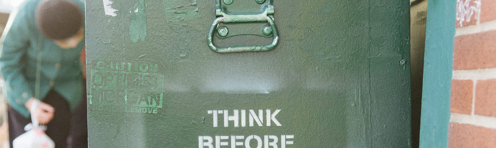 A green box book cart sitting outside a brick-and-mortar shop. Novels are piled on top of it. Across the side of the box, the words “Think before you speak. Read before you think.” are stenciled on in white letters.