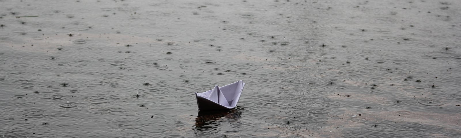 Photo of raindrops falling on water while a small paper boat bobs up and down.