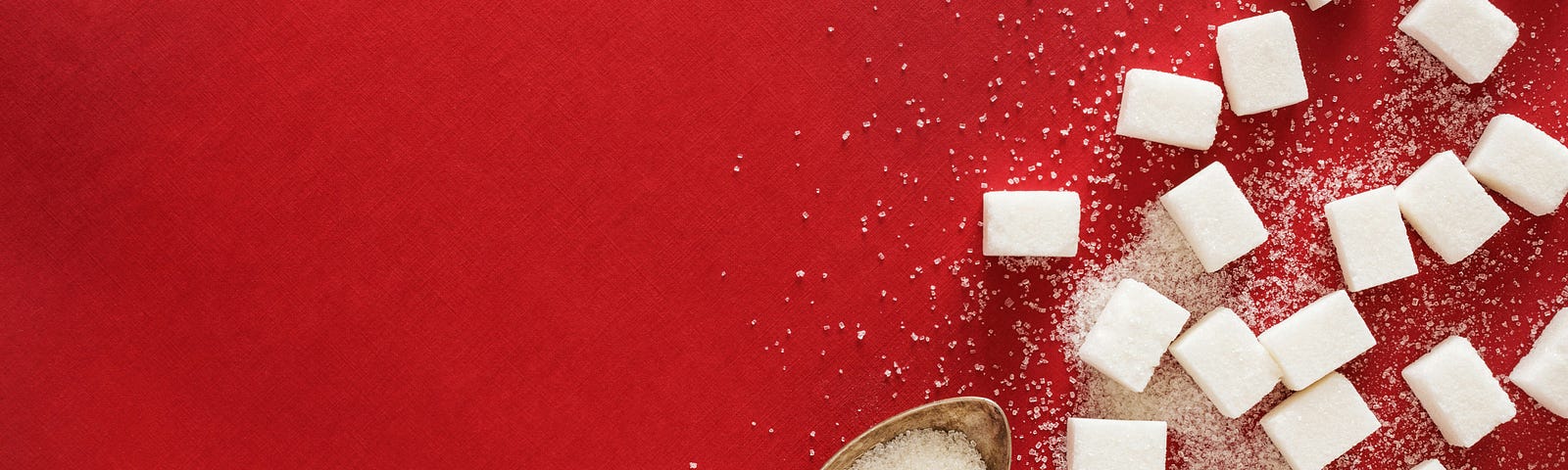 Sugar cubes scattered on a red background alongside a teaspoon of sugar.