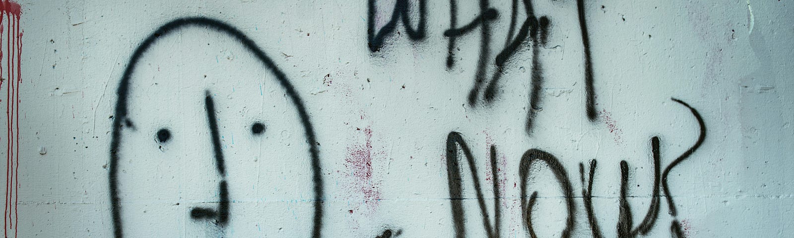 Graffiti of a black spray-painted face with the words ‘What Now’ scrawled on a roughly painted white wall.