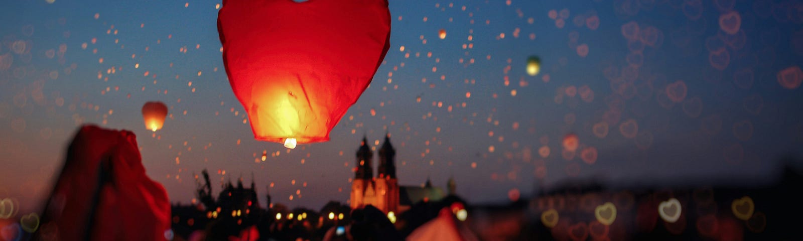 Red heart-shaped balloons being released against a dark blue night sky by a group of people, with one couple on a stage as if being honored.