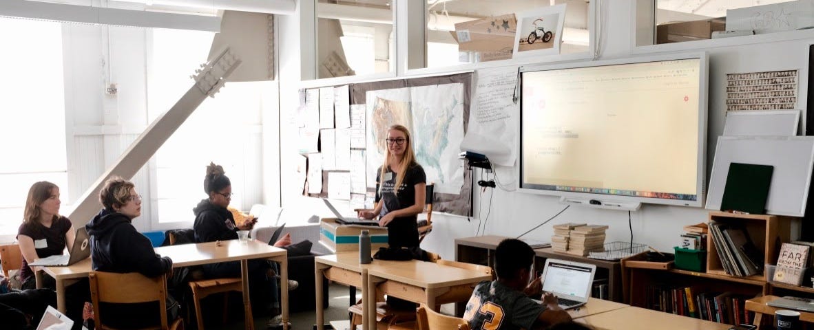 A young woman stands in front of the classroom looking at the camera. There are about six students around her.