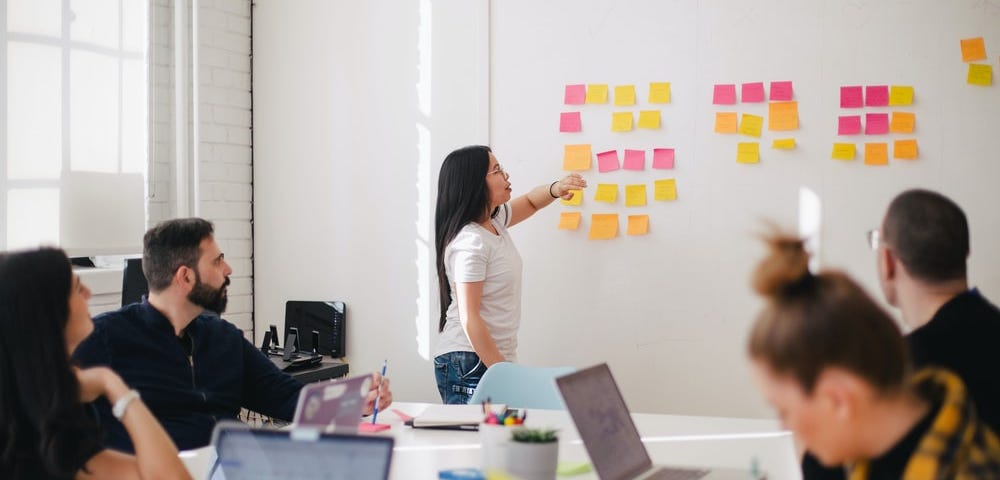 Grupo de colaboradores sentados, reunidos em uma sala branca em que há uma mulher em frente ao quadro de tarefas físico cheio de post-its representando cada tarefa.