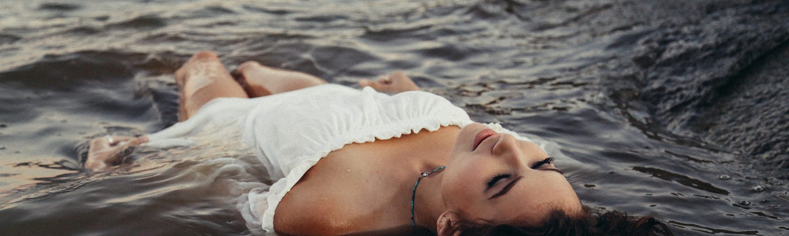 Glamorous woman in a white off the shoulder dress lying in beach water face up.